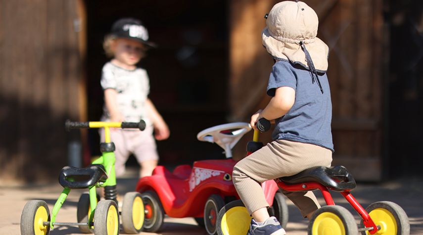 Ein Kind mit Sonnenhut sitzt auf einem roten Dreirad, während ein weiteres Kind mit Baseballkappe im Hintergrund steht. Mehrere bunte Kinderfahrzeuge stehen auf einem sonnigen Hof.