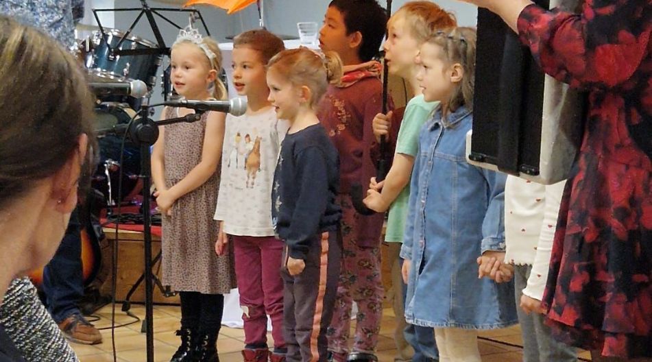 Eine Gruppe von Kindern steht auf einer Bühne und singt in Mikrofone. Im Hintergrund hält ein Kind einen orangen Regenschirm. Ein Erwachsener spielt ein Akkordeon, und einige Zuschauer sitzen im Vordergrund.