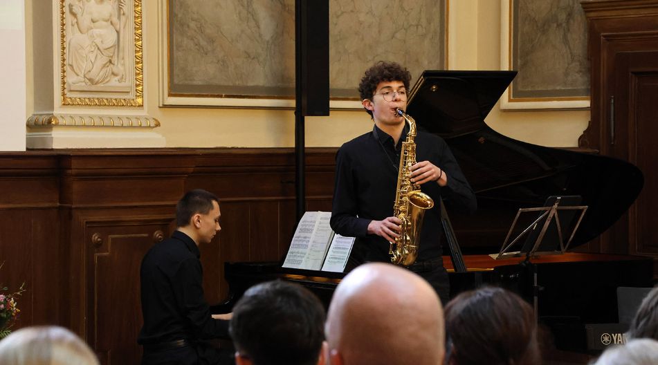 Ein Junger Mann steht und spielt auf einem Saxophon. Dahinter spielt ein weiterer junger Mann sitzend an einem Flügel.