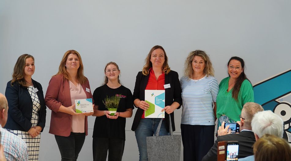 Sechs Frauen stehen bei einer Auszeichnungsveranstaltung des Berufswahl-Siegels nebeneinander und halten Urkunden, während Publikum fotografiert.