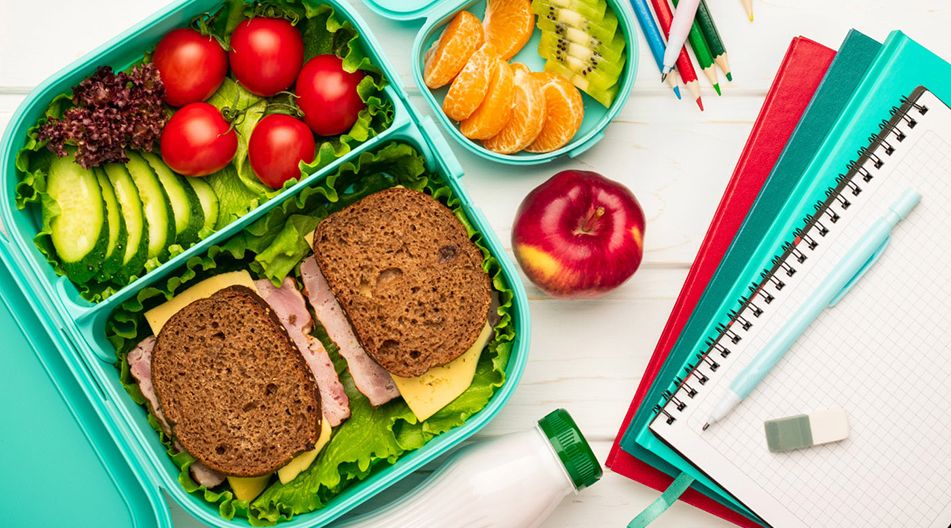 Geöffnete Frühstücksbox mit Vollkornbrot, Salat, Tomaten, Gurkenscheiben, daneben Schale mit Mandarinen- und Kiwistücken, Apfel, Trinkflasche, Notizbuch, Stifte und Radiergummi auf hellem Untergrund
