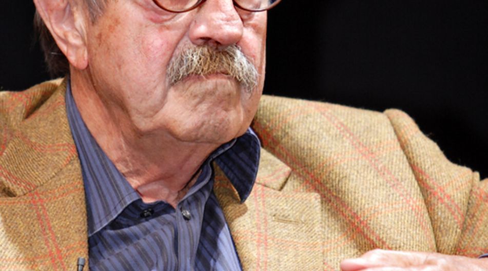 Älterer Mann (Günter Grass) mit Brille und Schnurrbart sitzt mit gefalteten Händen auf einem Sofa und blickt aufmerksam zur Seite.