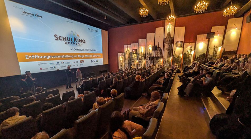 Kinosaal mit Publikum und zwei Personen auf der Bühne vor einer Leinwand mit der Aufschrift 'Schulkino Wochen'