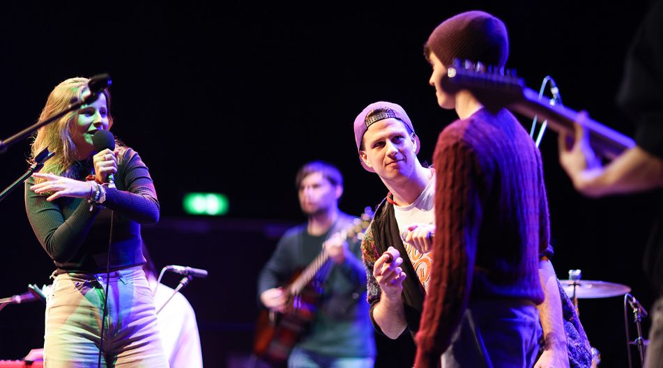Junge Musikerinnen und Musiker stehen gemeinsam auf einer Bühne; eine Frau singt ins Mikrofon, während andere mit Gitarre und Schlagzeug spielen.