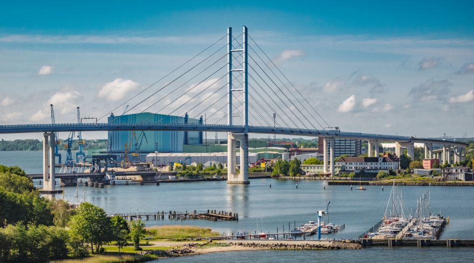 Blick auf die Rügenbrücke über das Wasser mit Hafen, Booten und Industrieanlagen im Hintergrund bei klarem Himmel.
