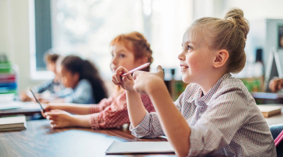 Kinder sitzen aufmerksam in einem Klassenzimmer; im Vordergrund ein blondes Mädchen mit Dutt, das lächelnd einen Stift in der Hand hält. Kinder sitzen aufmerksam in einem Klassenzimmer; im Vordergrund ein blondes Mädchen mit Dutt, das lächelnd einen Stift in der Hand hält.