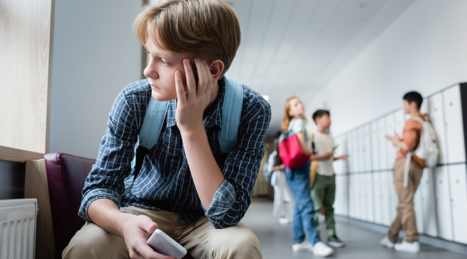 Junge sitzt allein auf einer Bank im Schulflur, wirkt nachdenklich, während andere Kinder im Hintergrund stehen.